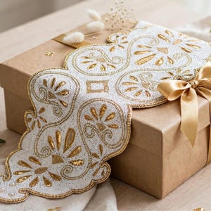 May include: A white and gold beaded table runner with a scalloped edge and intricate floral patterns, draped over a brown cardboard gift box tied with a smooth gold satin ribbon. The runner features dense white beadwork and gold accents.