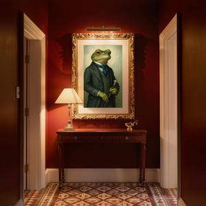 May include: A framed portrait of a green frog dressed in a dark grey suit, a white collared shirt, and a blue necktie. The frog holds a silver-topped walking cane. The artwork is displayed in a gold ornate frame on a deep red wall.