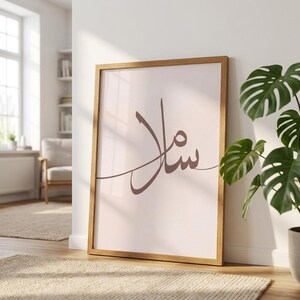 Peut inclure: Une affiche murale rectangulaire dans un cadre en bois clair présentant un fond rose pâle avec une calligraphie arabe brun foncé centrée. L'écriture se compose de lignes fluides et connectées formant un mot unique dans une teinte sombre.