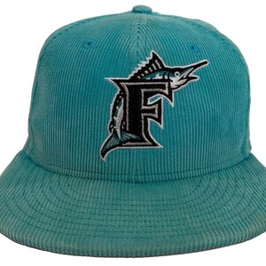 May include: A teal corduroy baseball cap featuring an embroidered logo on the front. The logo consists of a large black letter F with a white outline, overlaid with a detailed white and black graphic of a marlin fish jumping behind the letter.