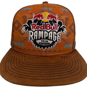 Op de afbeelding: Een bruine honkbalpet met een geometrisch patroon in oranje en lichtblauw. De voorkant toont een geborduurd Red Bull Rampage 11th Edition logo met twee rode stieren, een gele zon en witte tekst op een zwarte achtergrond met gekartelde randen.