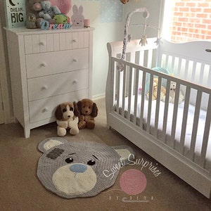 May include: A nursery with a white cot and chest of drawers, featuring a grey bear rug with a blue nose and a grey patch. Two soft toy dogs sit near the rug. A sign reads "Dream Big". A mobile hangs above the cot.