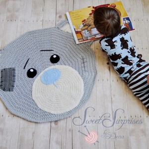May include: A grey crochet rug in the shape of a teddy bear's head. It has a white muzzle, blue nose, and black eyes. A child in blue bear-print pyjamas is lying beside the rug, reading a book. The rug is likely 100 cm in diameter.