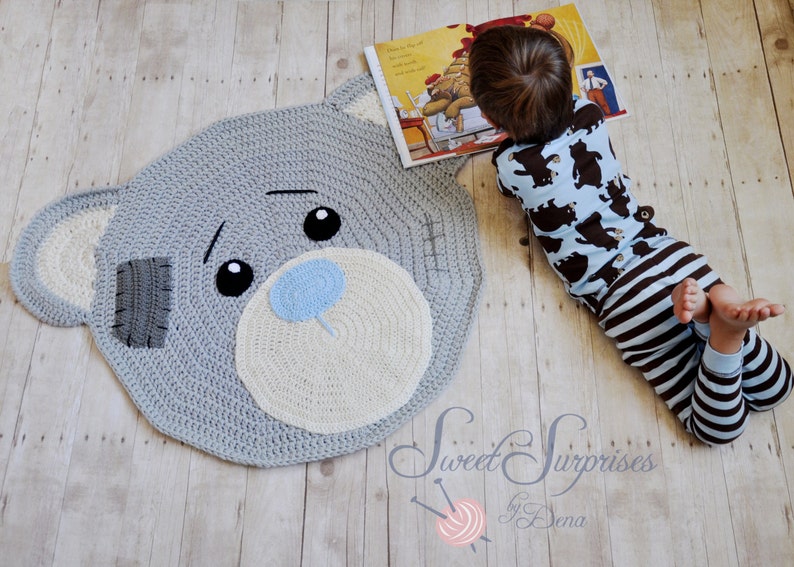 May include: A grey crochet rug in the shape of a teddy bear's head. It has a white muzzle, blue nose, and black eyes. A child in blue bear-print pyjamas is lying beside the rug, reading a book. The rug is likely 100 cm in diameter.