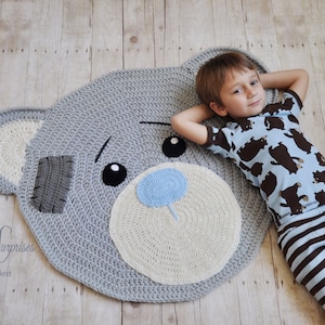 May include: A handmade, grey crochet rug in the shape of a teddy bear's face. The rug has a light blue nose, black eyes, and a beige muzzle. A grey patch is sewn on the bear's ear. The rug is on a wooden floor.