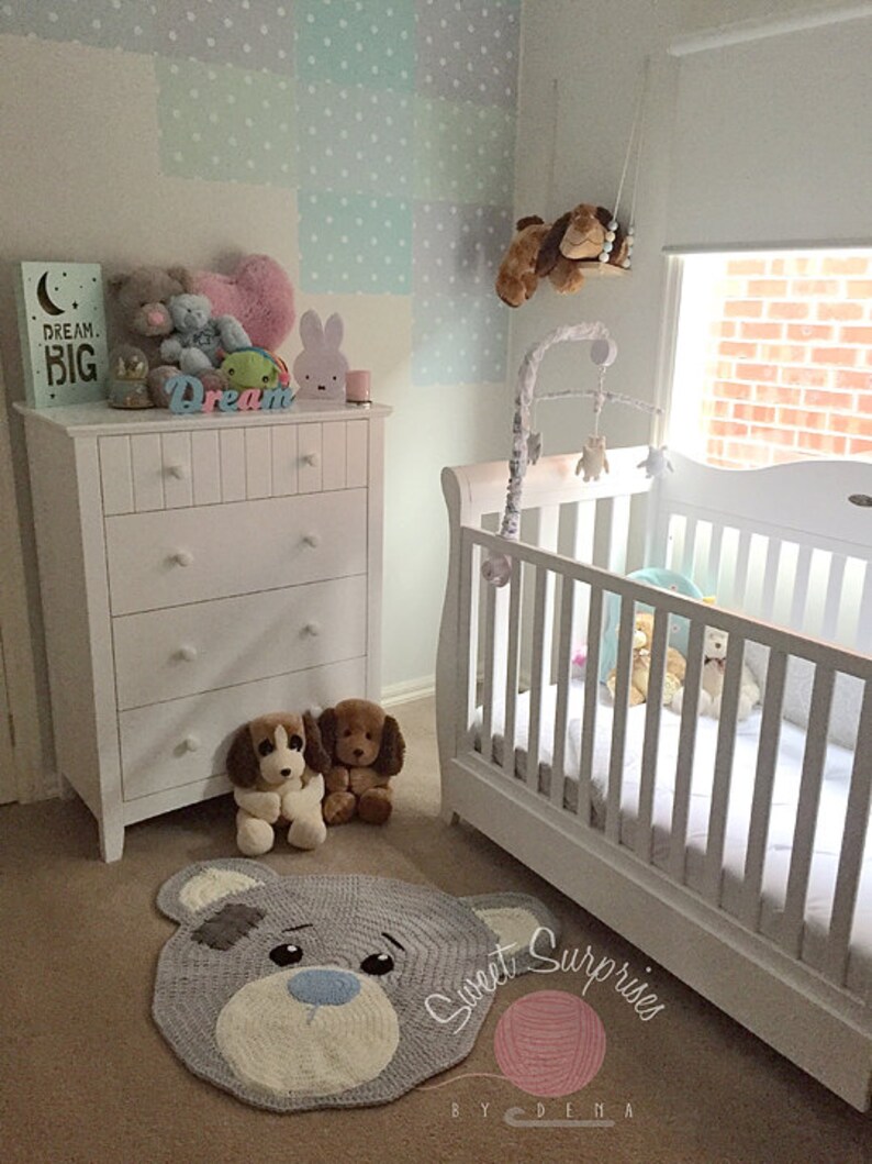 May include: A nursery with a white cot and chest of drawers, featuring a grey bear rug with a blue nose and a grey patch. Two soft toy dogs sit near the rug. A sign reads "Dream Big". A mobile hangs above the cot.