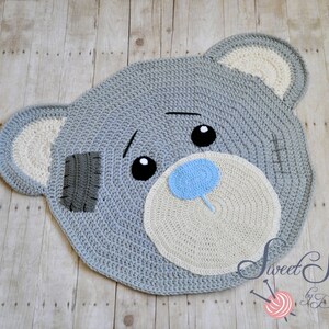May include: A handmade, grey crochet rug in the shape of a teddy bear's head. The rug features a light grey face, white ears, and a light blue nose. The bear has black eyes and a grey patch on its face. The rug is on a wooden surface.