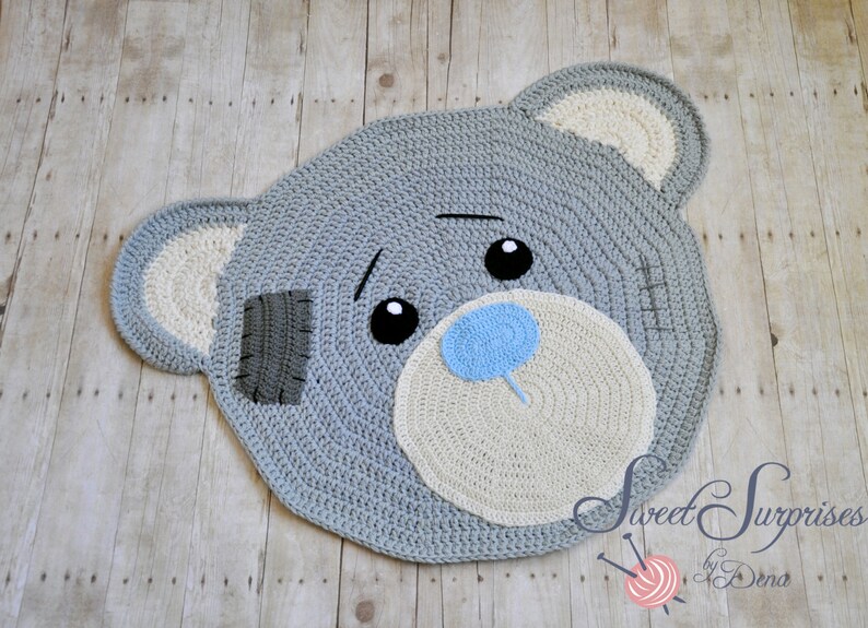 May include: A handmade, grey crochet rug in the shape of a teddy bear's head. The rug features a light grey face, white ears, and a light blue nose. The bear has black eyes and a grey patch on its face. The rug is on a wooden surface.