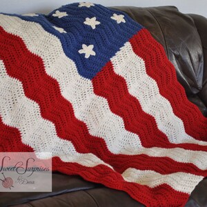 May include: A crocheted blanket in the style of the American flag, featuring red and white stripes and a blue field with white stars. The blanket is draped over a brown leather couch. The blanket is handmade.
