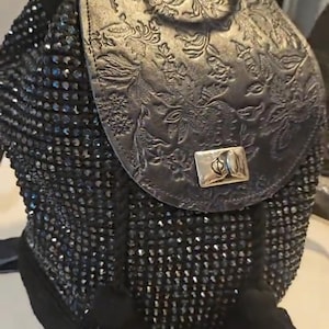 Peut inclure: Un sac à dos noir avec un panneau avant recouvert de petits cristaux noirs réfléchissants. Le rabat supérieur est en cuir noir avec un motif floral en relief et une fermeture à tourniquet en métal argenté. Deux pompons noirs pendent à l'avant.