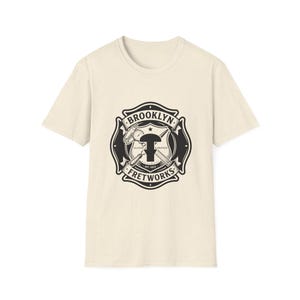 Puede incluir: Una camiseta de manga corta color crema con un gráfico circular negro en el pecho. El diseño incluye el texto BROOKLYN FRETWORKS, GUITAR REPAIR y EST. 2023, rodeando un icono central de martillo de trastes y herramientas cruzadas.
