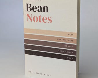 Może przedstawiać: Kremowy notatnik do kawy zatytułowany Bean Notes z gradientem pięciu poziomych brązowych pasków oznaczonych jako light, medium-light, medium, medium-dark i dark. Okładka zawiera tekst shops, beans, brews oraz 40 pp.