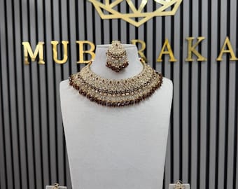 May include: A jewellery set consisting of a wide, ornate choker necklace, a matching maang tikka, and two dangling earrings. The pieces feature gold-tone metal, clear crystals, and dark brown teardrop-shaped beads arranged in a layered design.