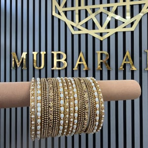 Op de afbeelding: Een set goudkleurige armbanden met afwisselende ontwerpen van kleine witte ovale kralen en getextureerde metalen banden. De collectie bevat verschillende individuele armbanden die op elkaar zijn gestapeld, met ingewikkelde patronen en een gepolijste afwerking.