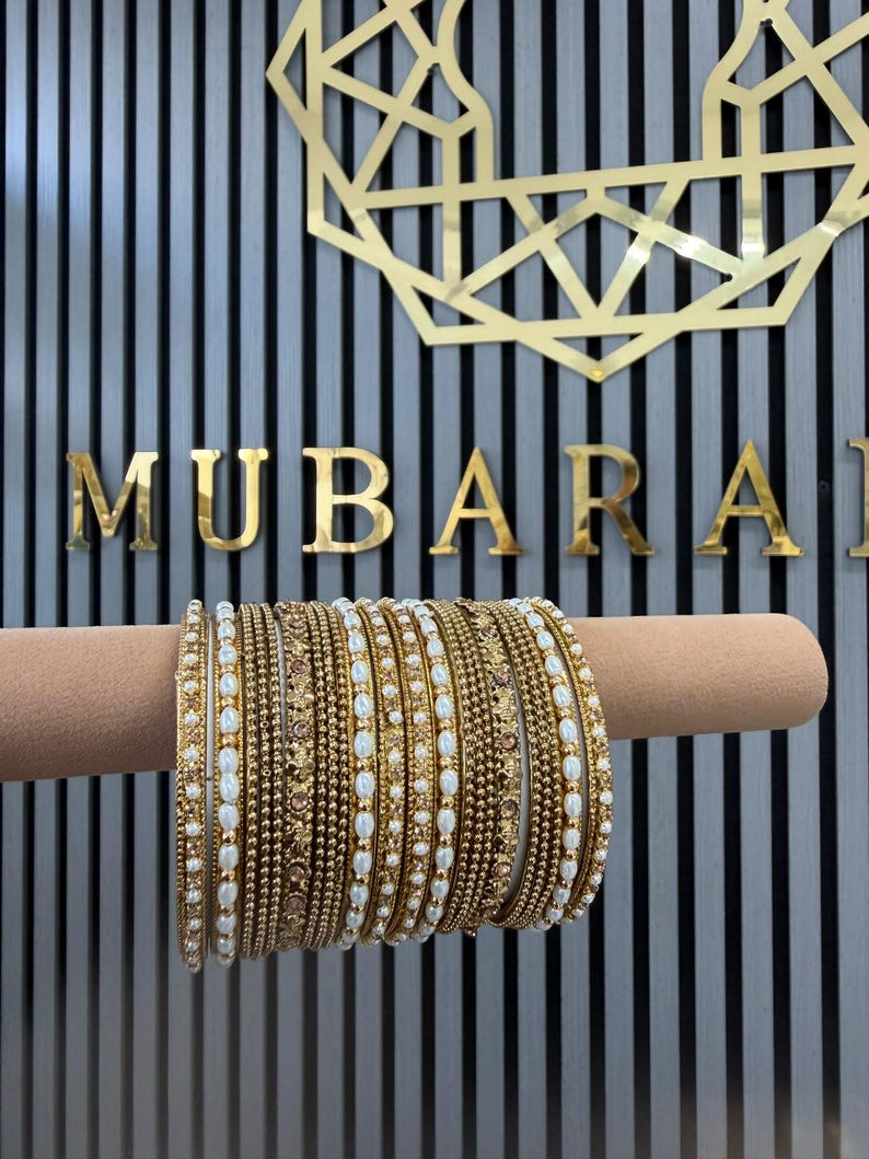 May include: A set of gold-toned bangles featuring alternating designs of small white oval beads and textured metallic bands. The collection includes several individual bracelets stacked together, showcasing intricate patterns and a polished finish.