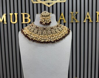 May include: A jewellery set featuring a wide, ornate choker necklace, a matching maang tikka, and two teardrop-shaped earrings. The pieces are crafted with antique gold-tone metal, embedded with dark red stones, and accented with cream-coloured pearls.