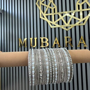 May include: A set of silver-toned bangles featuring alternating rows of small white faux pearls and clear sparkling crystals. The bracelets are arranged in a dense stack, showcasing a metallic finish with intricate beadwork and stone settings.