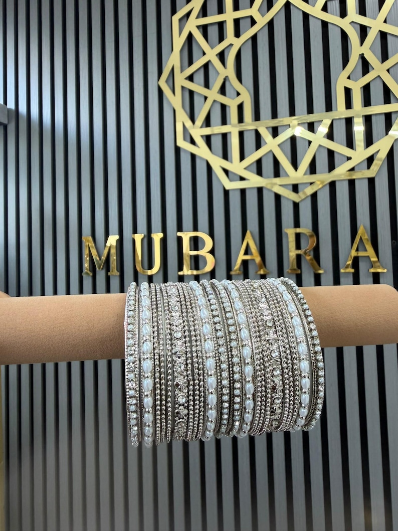 May include: A set of silver-toned bangles featuring alternating rows of small white faux pearls and clear sparkling crystals. The bracelets are arranged in a dense stack, showcasing a metallic finish with intricate beadwork and stone settings.