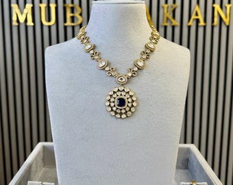 Könnte beinhalten: Ein goldfarbenes Halsketten- und Ohrringset mit einem zentralen, quadratischen, tiefblauen Stein, der von klaren Steinen in einem Blumenmuster umgeben ist. Die Kette besteht aus verbundenen goldfarbenen Segmenten mit klaren Steinen.