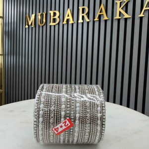 May include: A set of silver-toned bangles arranged in a stack, featuring intricate metallic beadwork, clear rhinestones, and white faux pearl accents. A small rectangular red tag with the white text 2*4 is attached to the front of the stack.