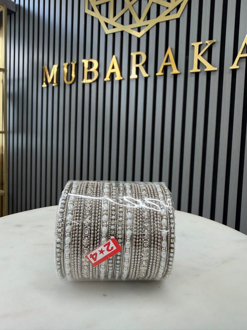 May include: A set of silver-toned bangles arranged in a stack, featuring intricate metallic beadwork, clear rhinestones, and white faux pearl accents. A small rectangular red tag with the white text 2*4 is attached to the front of the stack.