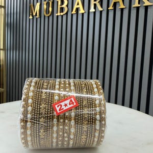 May include: A set of gold-toned metal bangles wrapped in clear plastic, featuring intricate textured patterns and embedded white faux pearls. A small red rectangular sticker with the white text 2*4 is attached to the front of the plastic wrap.