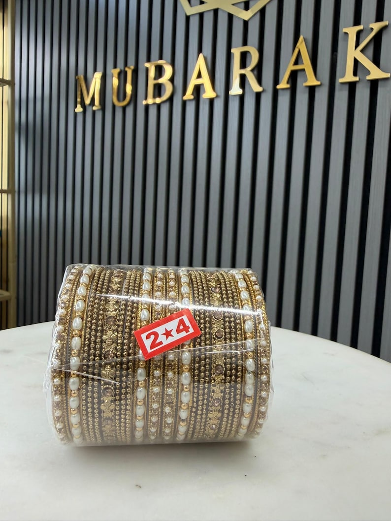 May include: A set of gold-toned metal bangles wrapped in clear plastic, featuring intricate textured patterns and embedded white faux pearls. A small red rectangular sticker with the white text 2*4 is attached to the front of the plastic wrap.