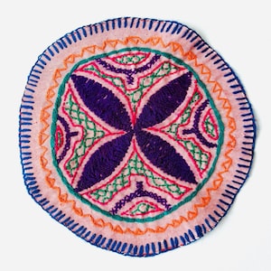 May include: A round, pink fabric patch with a colorful embroidered design. The design features a central motif of four purple petals with green and orange accents.