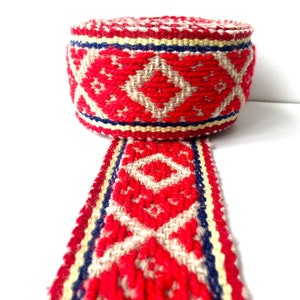 Puede incluir: Un rollo de cinta tejida roja con un patrón geométrico de diamantes en beige y blanco, acentuado con rayas azules y amarillas. La cinta se muestra sobre un fondo blanco, mostrando su intrincado diseño.