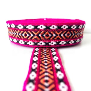 Puede incluir: Una cinta tejida rosa con un patrón geométrico negro, blanco y naranja. La cinta está enrollada y el patrón es visible en el lateral.