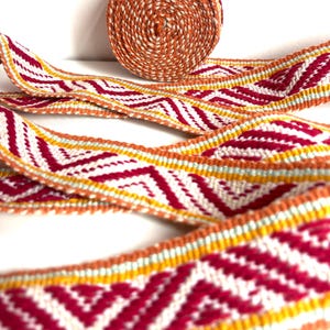Puede incluir: Un rollo de cinta de tela tejida con un patrón geométrico repetitivo de chevrón en tonos vibrantes de magenta, naranja brillante, blanco y verde menta pálido. La cinta textil está enrollada en la parte superior con una larga extensión.