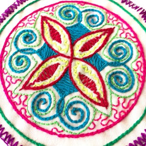 Puede incluir: Una pieza de tela circular con intrincados bordados a mano en tonos vibrantes de magenta, turquesa, rojo y amarillo. El diseño incluye un motivo floral central de cuatro pétalos rodeado de espirales, rematado con un borde de puntada zigzag morada.