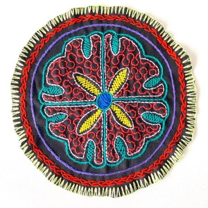 May include: A circular embroidered patch with a black background and a colorful floral design. The design features red, yellow, blue, and green thread. The patch is surrounded by a decorative border.