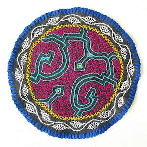 May include: A circular embroidered patch with a black background and a colorful geometric design. The design features a central motif of interlocking shapes in shades of pink, blue, and yellow, surrounded by a white and yellow border.