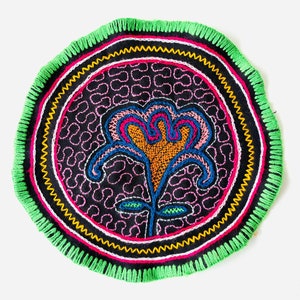May include: A circular, hand-stitched textile with a colorful floral design in the center. The flower is pink, orange, and blue, and the background is black with a repeating pattern. The textile is surrounded by a green border with a white and yellow trim.