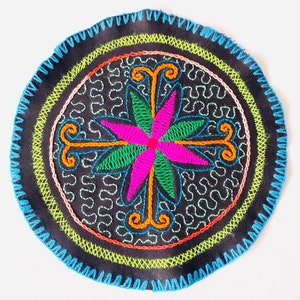 May include: A circular embroidered patch with a colorful floral design in the center. The design features a pink flower with green leaves and orange swirls. The patch is surrounded by a black background with a green, orange, and pink border.