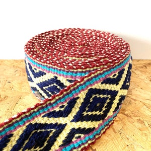 Puede incluir: Una cinta tejida con un patrón rojo, azul, amarillo y blanco. La cinta está enrollada y el patrón es visible en el exterior del rollo.