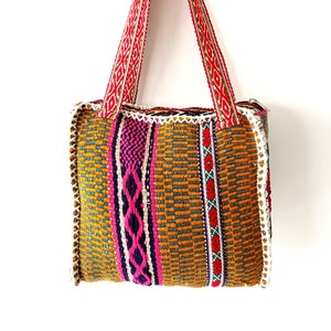 May include: A woven tote bag with a colorful geometric pattern in shades of orange, pink, green, and blue. The bag has a red and white woven strap.