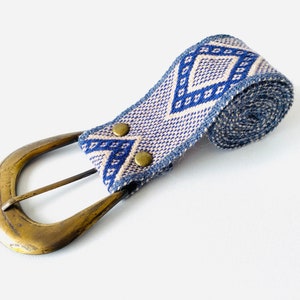 May include: A blue and white woven belt with a brass buckle. The belt is rolled up and the buckle is attached to the end of the belt.