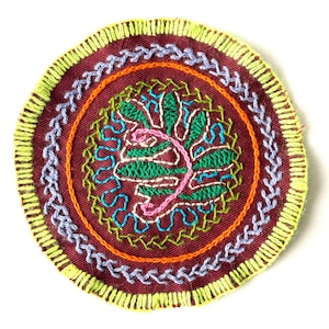 May include: A round, embroidered patch with a colorful floral design in the center. The patch is surrounded by multiple layers of stitching in shades of green, orange, purple, and yellow.