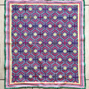May include: A square, colorful textile with a geometric pattern. The pattern is made up of repeating shapes in shades of pink, purple, blue, green, and yellow. The textile has a green border.