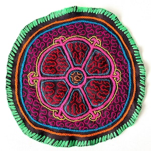 May include: A circular, hand-stitched fabric piece with a colorful, geometric design. The design features a central flower shape with pink, red, yellow, and blue embroidery on a black background. The fabric is edged with a green, orange, and blue trim.