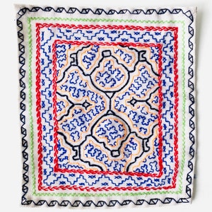 May include: A white fabric square with a colorful embroidered design. The design features a repeating pattern of blue, orange, and red shapes within a black outline. The embroidery is surrounded by a green, red, and black border.