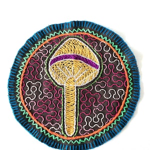 May include: A circular embroidered patch with a colorful design. The center of the patch features a yellow mushroom with a purple band around the top. The background is a dark brown fabric with a repeating pattern of white and pink shapes.