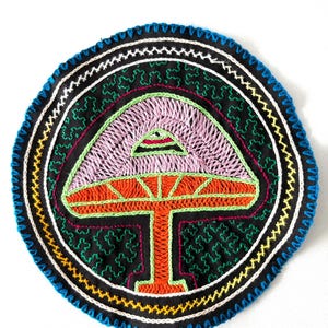 May include: A circular, black fabric patch with colorful embroidery depicting a stylized mushroom. The mushroom is outlined in orange and red, with a green stem and a pink cap. The fabric is surrounded by a blue, yellow, and white border.