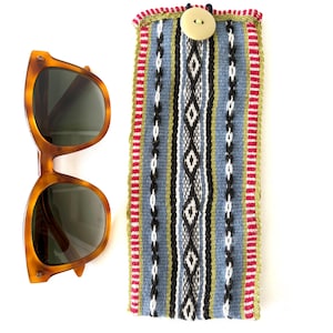 Puede incluir: Un estuche para gafas de sol de tela azul y verde con estampado y cierre de botón. El estuche está hecho de tela tejida con un borde rojo y blanco. El estuche está sobre una superficie blanca.