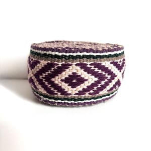 Puede incluir: Una banda textil tejida de forma cilíndrica con un patrón geométrico repetitivo de rombos en color morado oscuro, crema y verde oscuro. La tela presenta un tejido grueso y texturizado con rayas horizontales visibles.