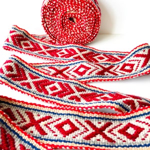 Puede incluir: Un rollo de cinta de tela tejida con un patrón geométrico repetitivo de diamantes en rojo vibrante, gris claro y azul marino. La cinta textil muestra un tejido texturizado e intrincado con un esquema de color consistente.