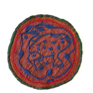 May include: Round, embroidered textile with a brown background and a blue frog design. The frog is surrounded by blue leaves and dots. The border features red, orange, and green stitching.