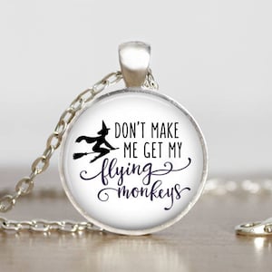 May include: A silver necklace with a round glass pendant featuring a black silhouette of a witch flying on a broomstick. The pendant has the text "Don't make me get my flying monkeys".